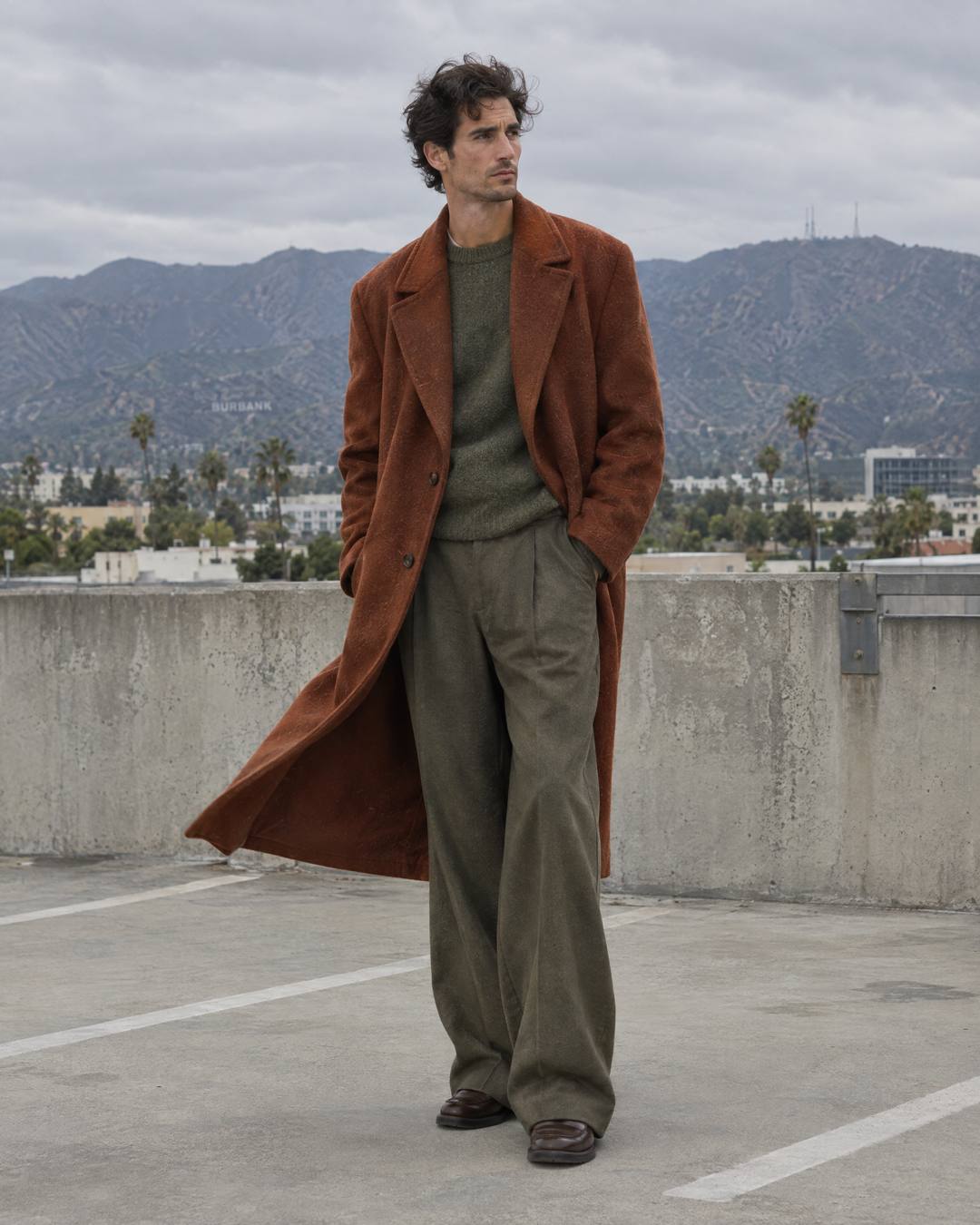 Man in a rust wool overcoat on a rooftop parking structure with mountains behind